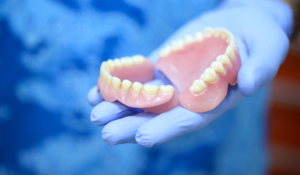 Image of someone in a blue doctor gown and blue latex gloves holding a set of dentures, possibly getting ready for a denture wash impression